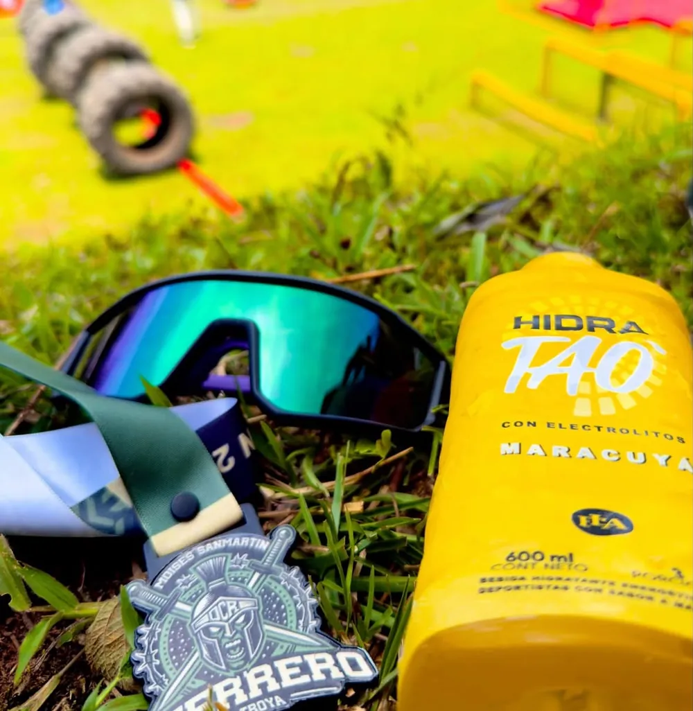 Medalla finisher de Guerrero de Troya junto a una botella de Hidratao y gafas deportivas, sobre el pasto del recorrido